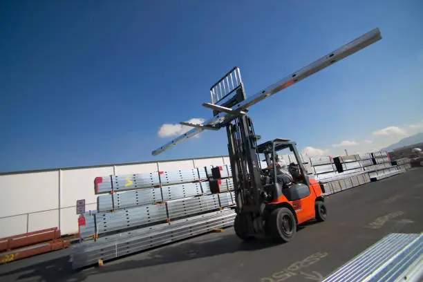 Forklift moving long metal beams outside warehouse.